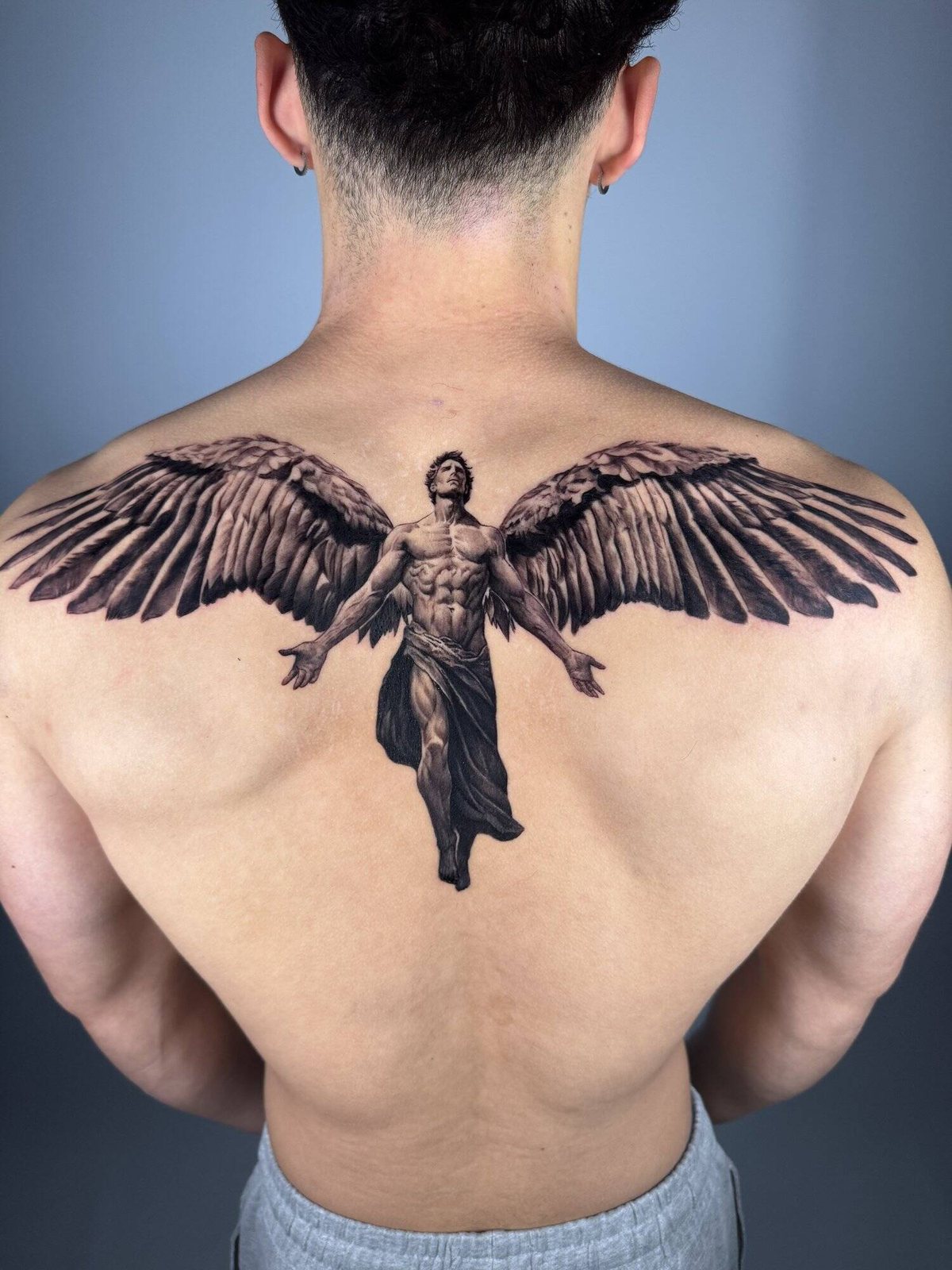 Black and grey realism upper back tattoo of a winged angel with outstretched wings spanning the shoulders