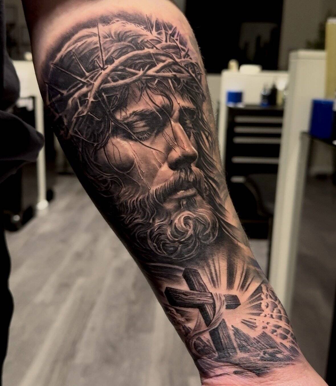Black and grey realism tattoo of Christ wearing the crown of thorns, running down a forearm