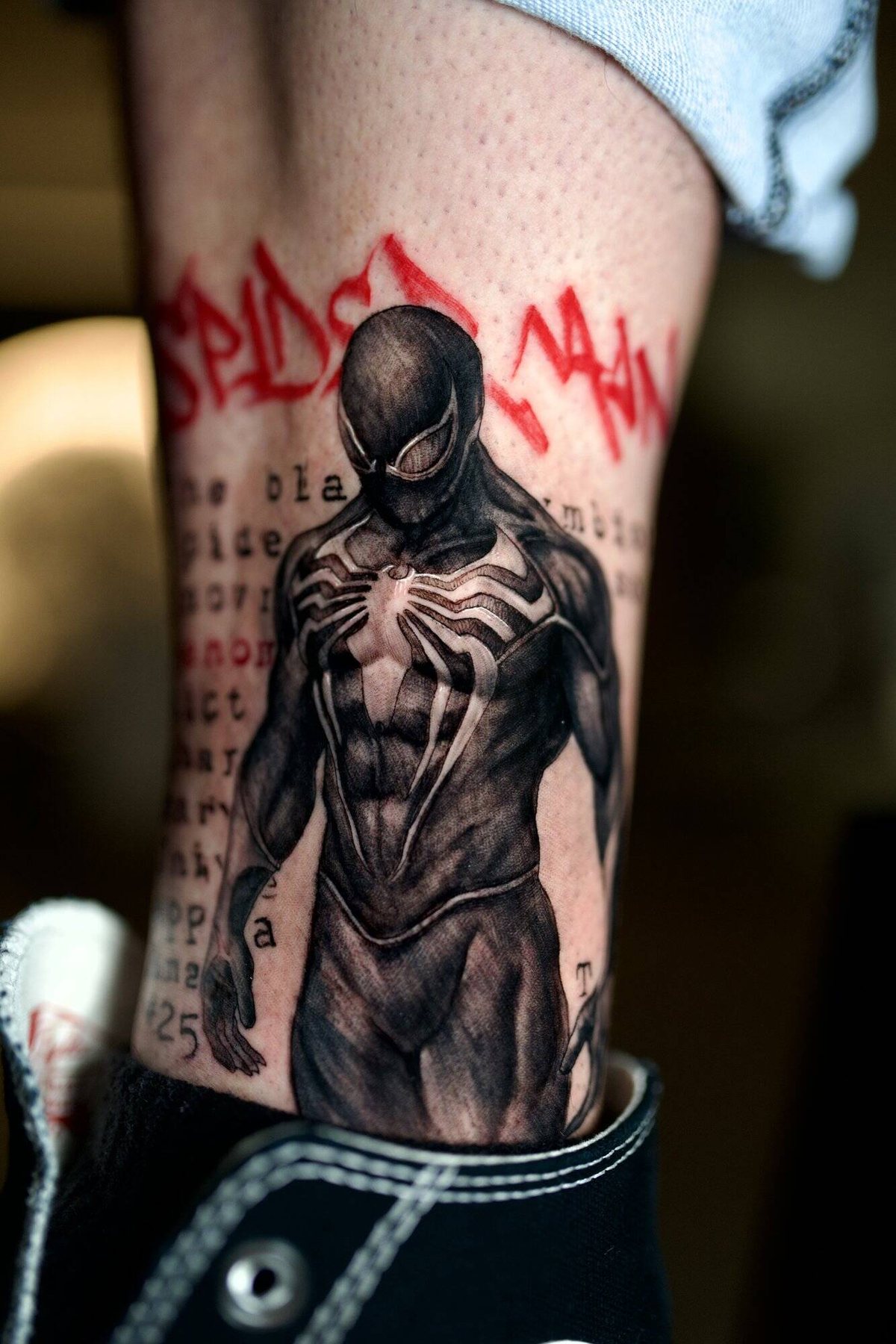 Black and grey realism calf tattoo of a symbiote Spider-Man figure with lettering behind it